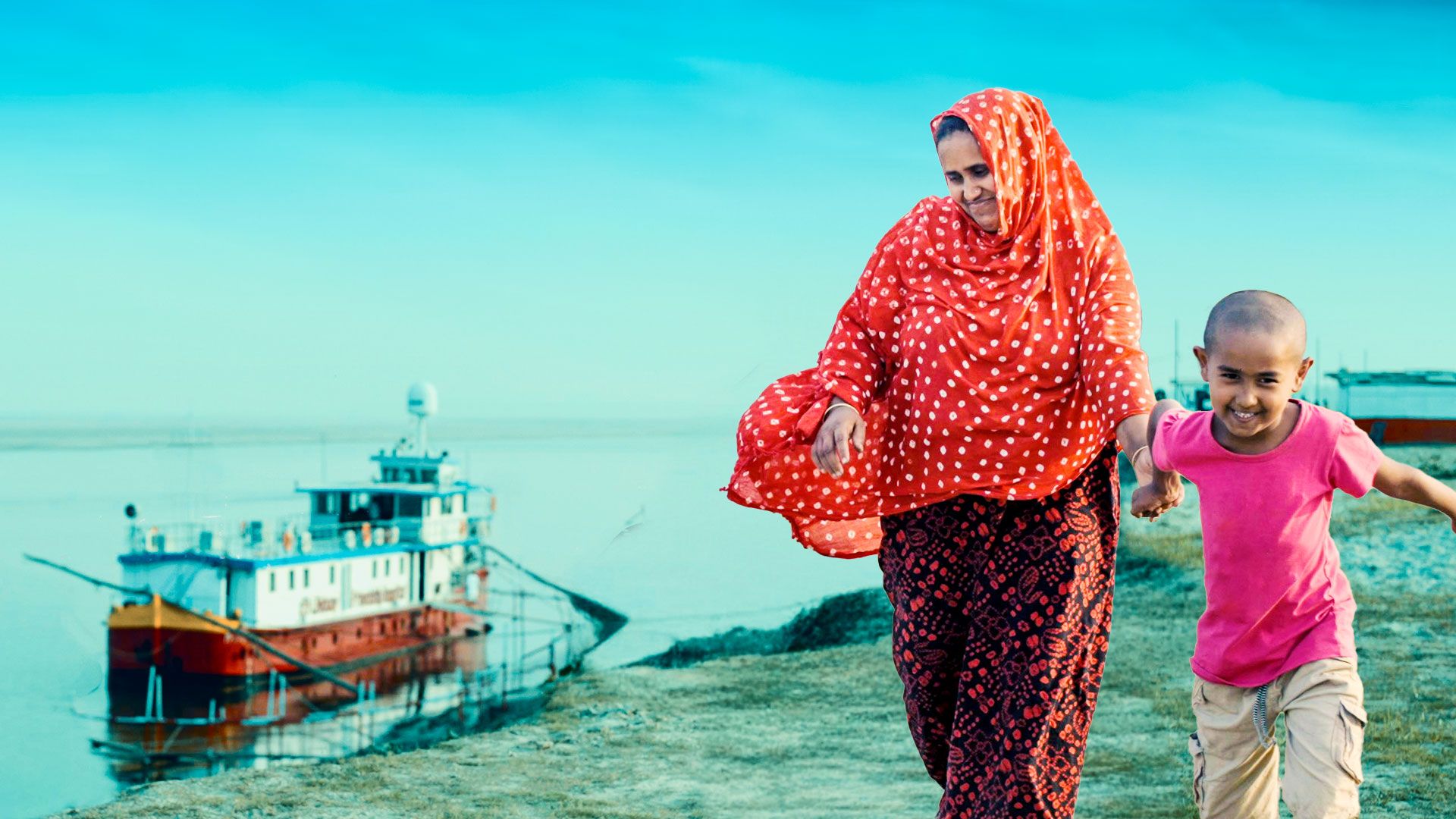 A mother and child from the char (river-island) area walk by, with the Lifebuoy Friendship Hospital in the background