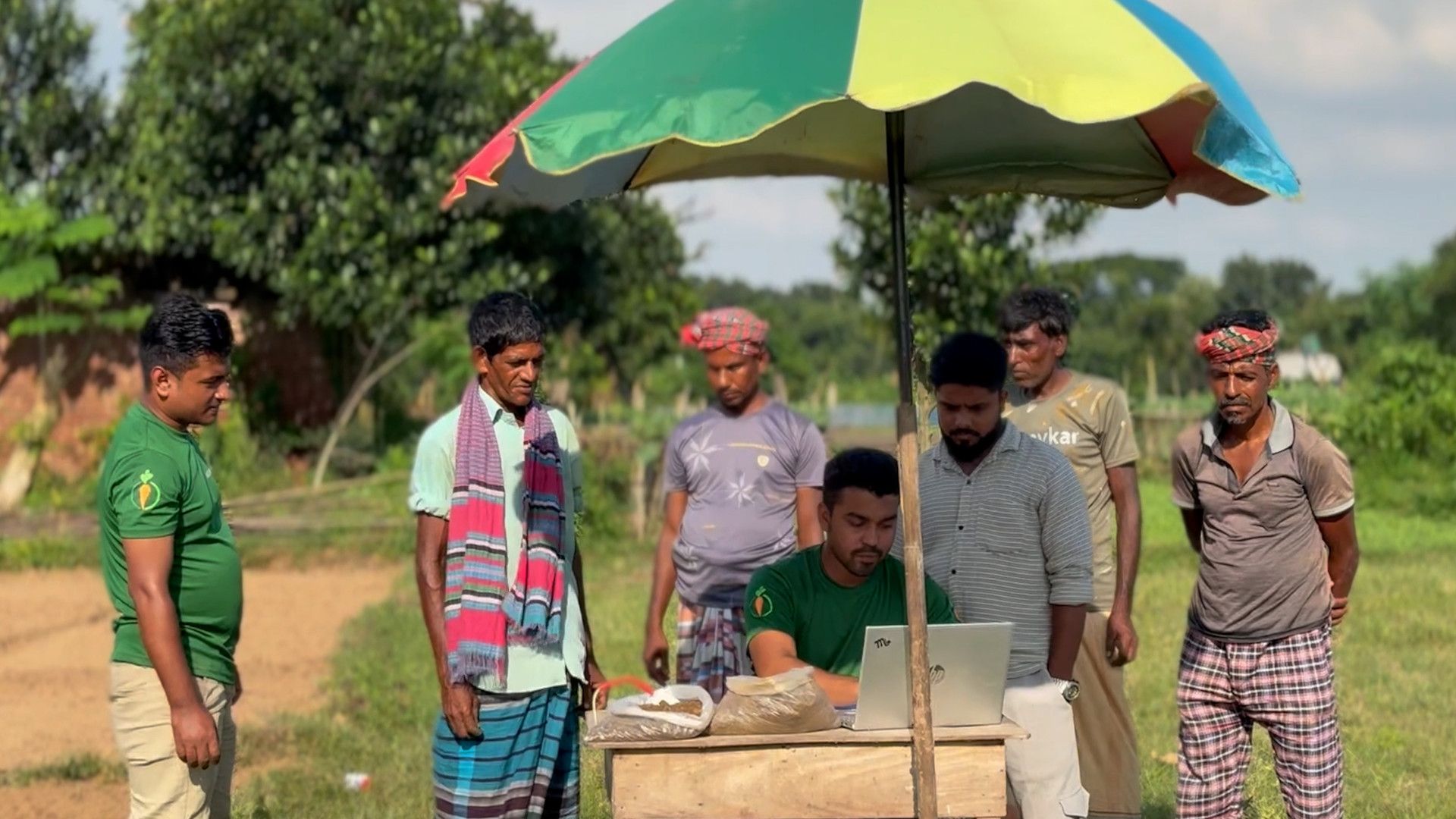 Field officer recording crop data with farmers under umbrella.